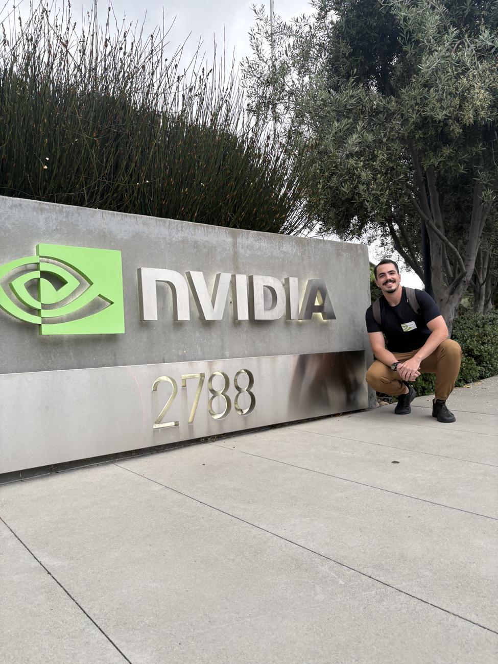 Matheus Duzzi agachado ao lado do letreiro da NVIDIA na sede em Santa Clara